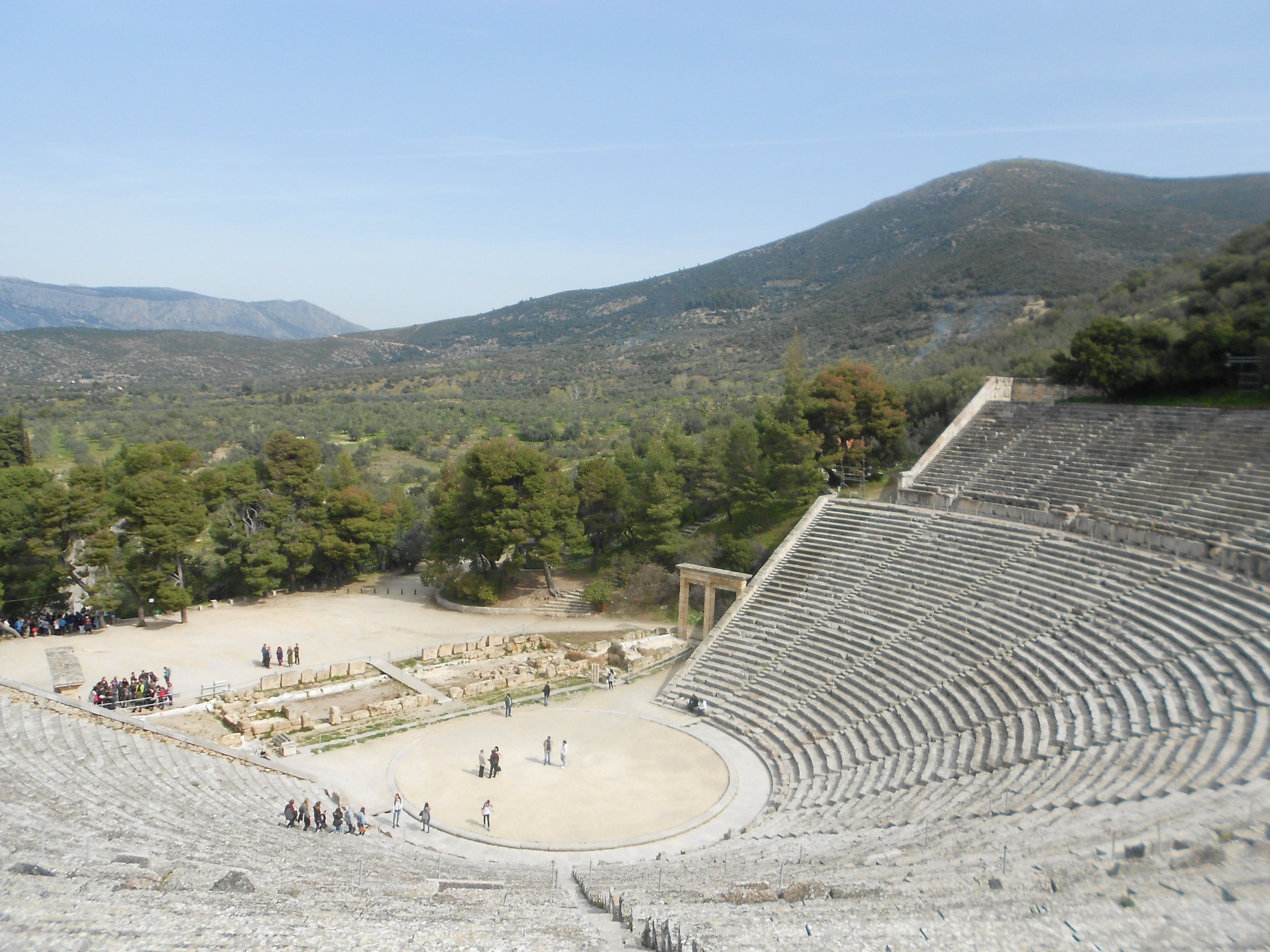 The ancient theatre of Epidaurus, tiers of stone seats curving above the circular orchestra with wooded hills beyond